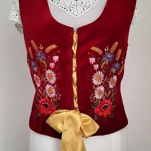 K&ouml;nnte beinhalten: Eine rote Samt-Weste mit einem goldenen Band und gestickten Blumenmustern. Die Weste hat einen Spitzenausschnitt und eine Schn&uuml;rung im Korsett-Stil an der Vorderseite.