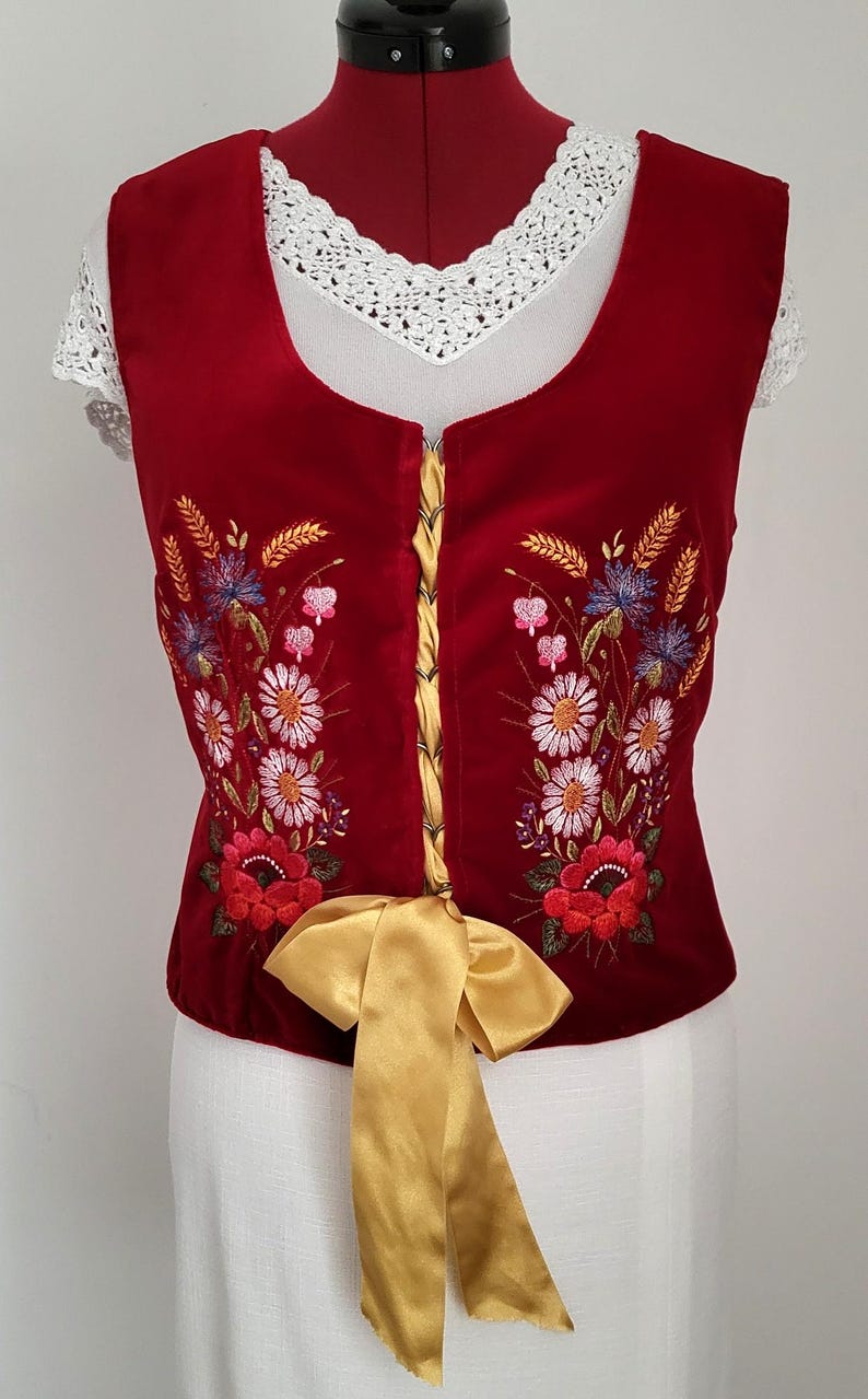 K&ouml;nnte beinhalten: Eine rote Samt-Weste mit einem goldenen Band und gestickten Blumenmustern. Die Weste hat einen Spitzenausschnitt und eine Schn&uuml;rung im Korsett-Stil an der Vorderseite.