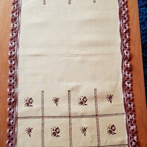 May include: A beige table runner with a brown crocheted border and a brown floral pattern. The runner is divided into squares with a brown line.