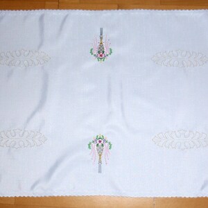 May include: White tablecloth with embroidered floral designs in shades of pink, green, and white. The tablecloth has a scalloped edge.