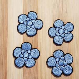 May include: Four blue embroidered flower patches with black outlines. The flowers have a five-petal design with a central circle.