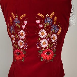 K&ouml;nnte beinhalten: Rote Samt-Weste mit gesticktem Blumendesign mit wei&szlig;en, roten, rosa, blauen und gelben Blumen. Die Weste ist &auml;rmellos und hat einen runden Ausschnitt.