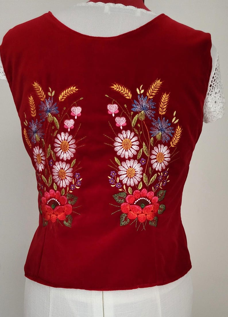 K&ouml;nnte beinhalten: Rote Samt-Weste mit gesticktem Blumendesign mit wei&szlig;en, roten, rosa, blauen und gelben Blumen. Die Weste ist &auml;rmellos und hat einen runden Ausschnitt.