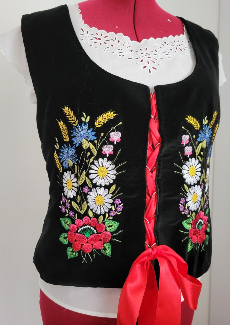 K&ouml;nnte beinhalten: Eine schwarze Samt-Weste mit roter Schleife und gestickten Blumendesigns in Rot-, Wei&szlig;-, Blau-, Gelb- und Gr&uuml;nt&ouml;nen.