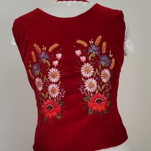 K&ouml;nnte beinhalten: Eine rote, &auml;rmellose Weste mit gestickten Blumenmustern. Die Blumen sind wei&szlig;, rosa, blau und orange. Die Weste hat einen runden Ausschnitt.