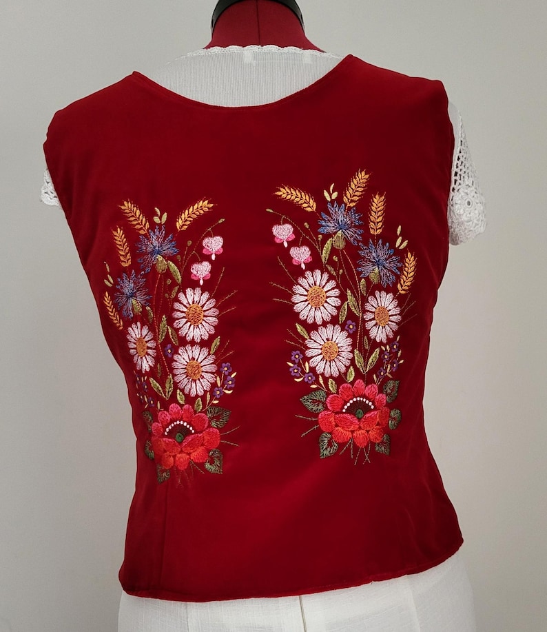 K&ouml;nnte beinhalten: Eine rote, &auml;rmellose Weste mit gestickten Blumenmustern. Die Blumen sind wei&szlig;, rosa, blau und orange. Die Weste hat einen runden Ausschnitt.
