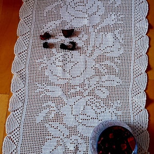 May include: White crocheted table runner with a floral design. The runner is on a wooden surface with a glass bowl filled with red potpourri.