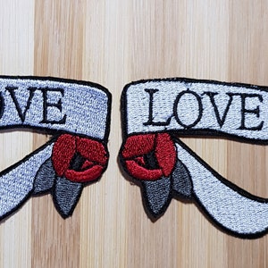 Puede incluir: Dos parches bordados con la palabra "LOVE" en una pancarta blanca con una rosa roja a cada lado.