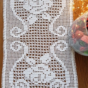 May include: White crocheted table runner with a delicate floral pattern. The runner is made of fine yarn and has a lacy texture.