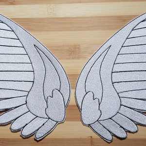 May include: Two embroidered, symmetrical white angel wing patches with black outline stitching. The wings are detailed with layered feather patterns, perfect for adding to clothing or accessories. The patches are displayed on a light brown wooden surface.