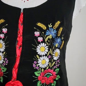 K&ouml;nnte beinhalten: Eine schwarze Samt-Weste mit einem roten Band und gestickten Blumen in den Farben Wei&szlig;, Gelb, Blau, Rosa und Gr&uuml;n.