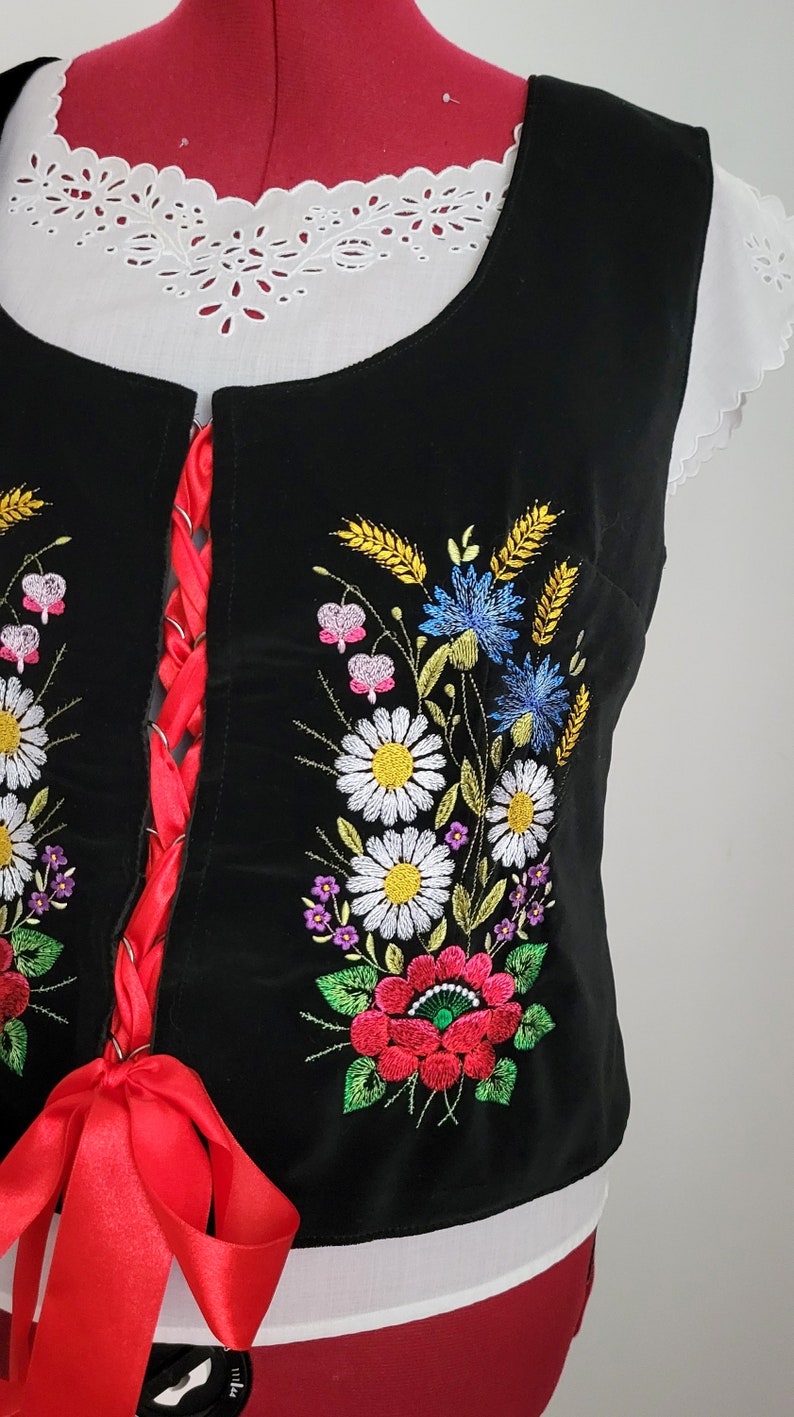 K&ouml;nnte beinhalten: Eine schwarze Samt-Weste mit einem roten Band und gestickten Blumen in den Farben Wei&szlig;, Gelb, Blau, Rosa und Gr&uuml;n.