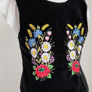 K&ouml;nnte beinhalten: Schwarze &auml;rmellose Weste mit gestickten Blumendesigns in Rot-, Gelb-, Wei&szlig;-, Blau- und Gr&uuml;nt&ouml;nen. Die Blumen sind in zwei symmetrischen B&uuml;scheln auf der Vorderseite der Weste angeordnet.