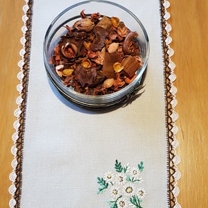 May include: White linen table runner with brown crocheted edging and embroidered white flowers. A glass bowl filled with potpourri sits in the centre of the runner.