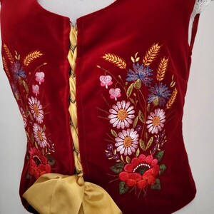 K&ouml;nnte beinhalten: Eine rote Samt-Weste mit einem goldenen Band, das in der Taille gebunden ist. Die Weste ist mit einem Blumendesign in Wei&szlig;-, Rosa-, Blau- und Gr&uuml;nt&ouml;nen bestickt.
