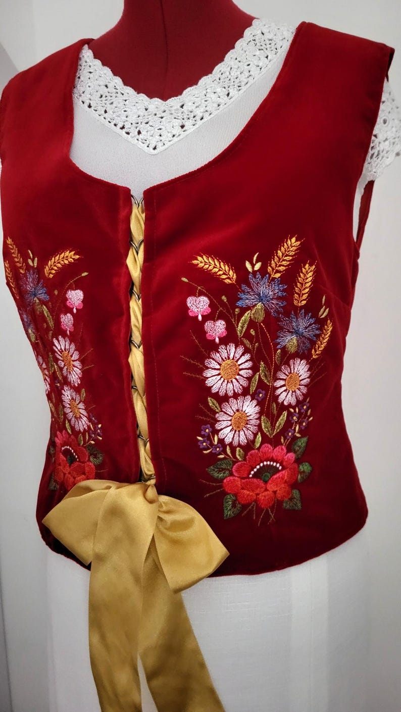 K&ouml;nnte beinhalten: Eine rote Samt-Weste mit einem goldenen Band, das in der Taille gebunden ist. Die Weste ist mit einem Blumendesign in Wei&szlig;-, Rosa-, Blau- und Gr&uuml;nt&ouml;nen bestickt.