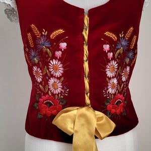 K&ouml;nnte beinhalten: Eine rote Samt-Weste mit gestickten Blumenmotiven in den Farben Rosa, Wei&szlig;, Blau und Gr&uuml;n. Die Weste hat ein goldenes Band, das an der Taille zu einer Schleife gebunden ist.