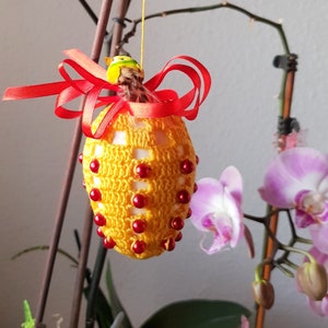 Op de afbeelding: Een geel gehaakt paasei met rode kralen en een rood lint. Het ei hangt aan een tak met roze orchideeën bloemen.
