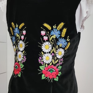 K&ouml;nnte beinhalten: Schwarze &auml;rmellose Weste mit gestickten Blumendesigns in Rot-, Gelb-, Blau-, Gr&uuml;n- und Wei&szlig;t&ouml;nen. Die Blumen umfassen G&auml;nsebl&uuml;mchen, Kornblumen und Weizen.