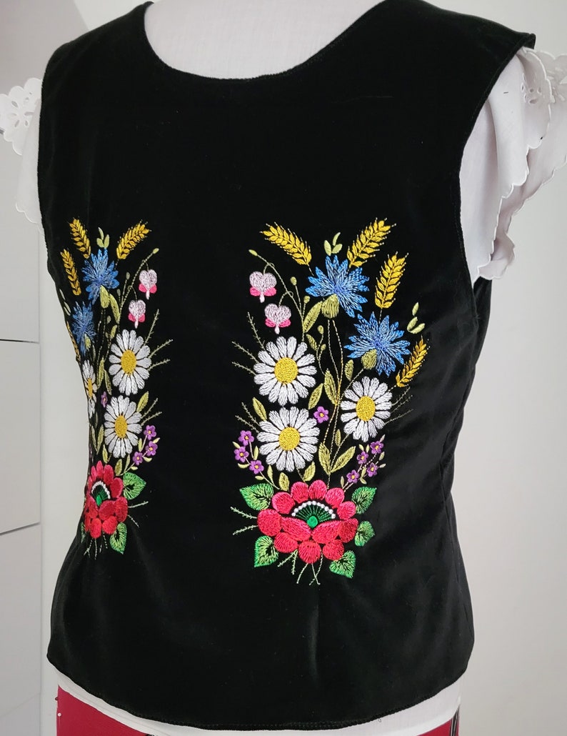 K&ouml;nnte beinhalten: Schwarze &auml;rmellose Weste mit gestickten Blumendesigns in Rot-, Gelb-, Blau-, Gr&uuml;n- und Wei&szlig;t&ouml;nen. Die Blumen umfassen G&auml;nsebl&uuml;mchen, Kornblumen und Weizen.