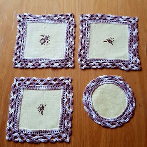 May include: Set of four crocheted doilies in white and purple with a light yellow linen center. Each doily features a floral design.