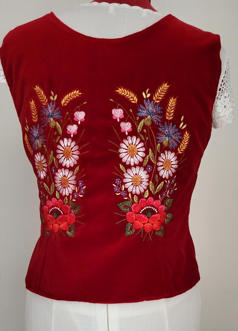 K&ouml;nnte beinhalten: Rote Samt-Weste mit gesticktem Blumendesign mit wei&szlig;en, roten, rosa, blauen und gelben Blumen. Die Weste ist &auml;rmellos und hat einen runden Ausschnitt.