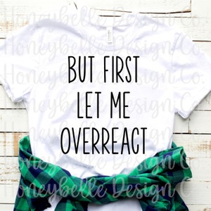 Puede incluir: Camiseta blanca con texto negro que dice "But first let me overreact". La camiseta está doblada y colocada sobre una superficie de madera con una camisa de cuadros verde y negro encima.