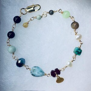 Pode incluir: Um bracelete de corrente dourada com uma variedade de pedras preciosas, incluindo preto, verde, azul e branco. O bracelete tem um fecho dourado e um pequeno amuleto dourado.