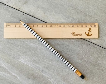 May include: A wooden ruler with a black and white striped pencil on top of it. The ruler is marked with centimetres and numbers from 0 to 20. The ruler has the name "Bame" and an anchor symbol engraved on it.
