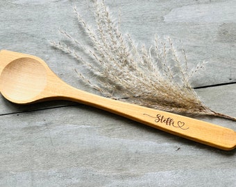 May include: A wooden spoon with a heart and the name "Steffi" engraved on the handle. The spoon is lying on a light gray wooden surface with dried grass behind it.