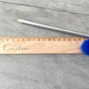 May include: A wooden ruler with a blue pom-pom attached to the end. The ruler is marked in centimetres and has the name "Emilia" engraved on it.