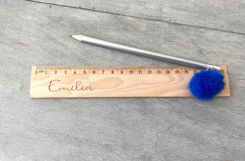 May include: A wooden ruler with a blue pom-pom attached to the end. The ruler is marked in centimetres and has the name "Emilia" engraved on it.