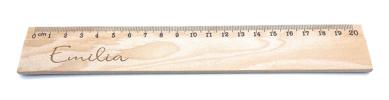 May include: A wooden ruler with a personalised inscription that reads "Emilia". The ruler is 20 centimetres long and has markings for each centimetre.