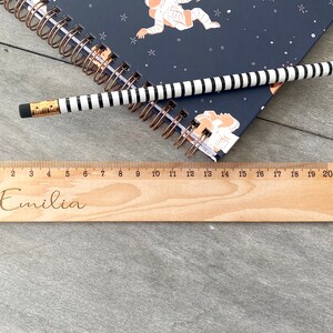 May include: A wooden ruler with a personalised inscription that reads "Emilia". The ruler is 20 centimetres long and has markings for each centimetre.