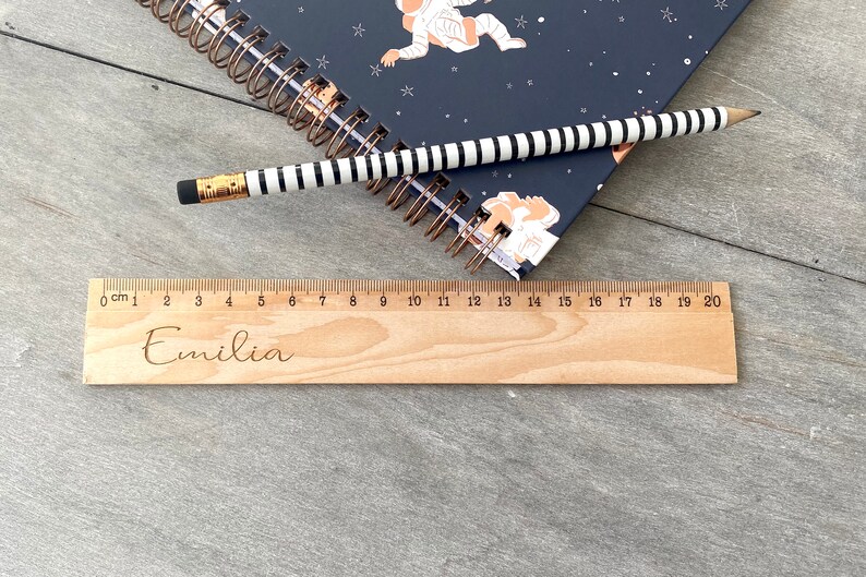 May include: A wooden ruler with a personalised inscription that reads "Emilia". The ruler is 20 centimetres long and has markings for each centimetre.