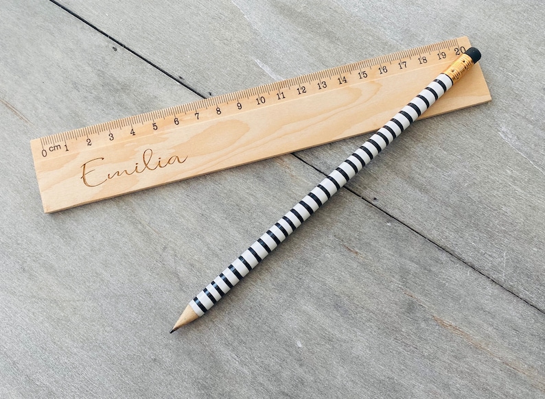 May include: A wooden ruler with black and white stripes and the name "Emilia" engraved on it. The ruler is 20 centimetres long and has markings for each centimetre.