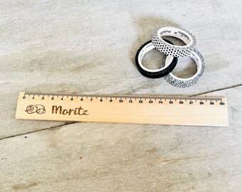 Könnte beinhalten: Ein Holzlineal mit einer personalisierten Inschrift, die "Moritz" lautet. Das Lineal ist 50,8 cm lang und hat schwarze Markierungen für jeden Zentimeter. Im Hintergrund befinden sich drei Rollen gemusterten Washi-Bands.