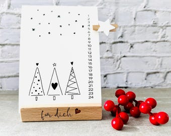 May include: A white countdown calendar with black numbers 1-24, a black and white illustration of three Christmas trees, and the text "fur dich" on a wooden base.