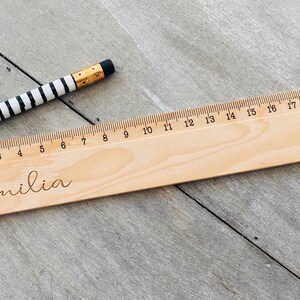 May include: A wooden ruler with a black and white striped pencil on top. The ruler is marked with centimetres and numbers from 0 to 20. The name "Emilia" is engraved on the ruler.