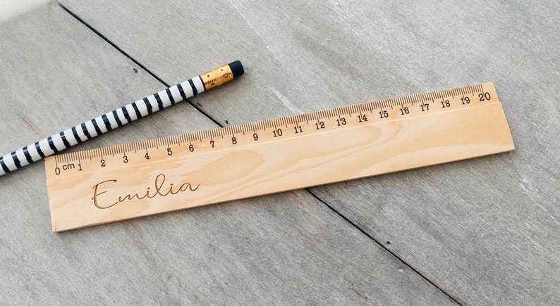 May include: A wooden ruler with a black and white striped pencil on top. The ruler is marked with centimetres and numbers from 0 to 20. The name "Emilia" is engraved on the ruler.