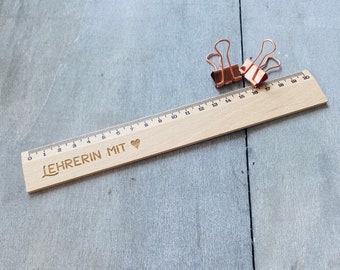 Könnte beinhalten: Ein Holzlineal mit einer schwarzen Aufschrift "LEHRERIN MIT" und einem Herzsymbol. Das Lineal ist 50 cm lang und hat schwarze Markierungen für jeden Zentimeter. Zwei goldene Büroklammern liegen auf dem Lineal.