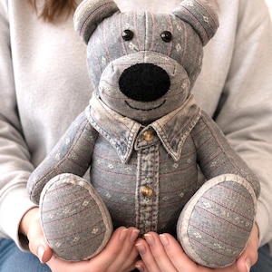 May include: A gray teddy bear made from patterned fabric, featuring a black nose and button eyes. The bear wears a denim shirt with two gold buttons. The bear is a soft toy.