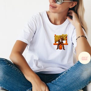 May include: White t-shirt with a graphic of a man in a suit sitting on a bench, with the words "Benito Bowl" in yellow and orange. The shirt is paired with blue jeans.