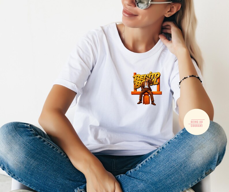 May include: White t-shirt with a graphic of a man in a suit sitting on a bench, with the words "Benito Bowl" in yellow and orange. The shirt is paired with blue jeans.