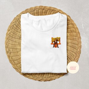 May include: White t-shirt with a small graphic of a man sitting on a bench with the words "Bento Box" in orange and yellow. The shirt is folded on a woven, circular mat.