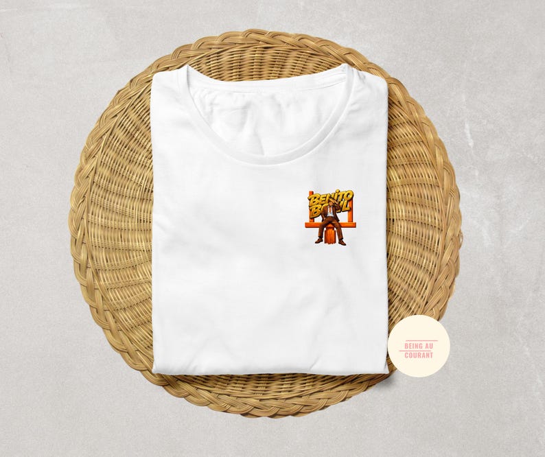 May include: White t-shirt with a small graphic of a man sitting on a bench with the words "Bento Box" in orange and yellow. The shirt is folded on a woven, circular mat.