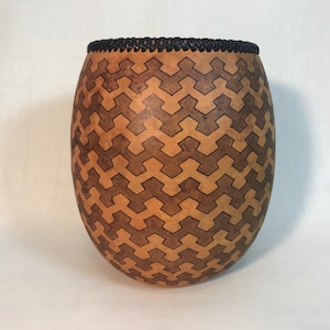 May include: A handcrafted gourd vase with a geometric pattern. The vase has a natural tan color with a repeating dark brown design. The top edge is finished with a black, woven trim.
