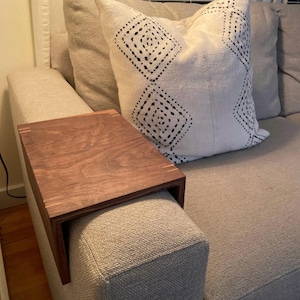 Puede incluir: Una bandeja de brazo de sofá de madera con un acabado marrón cálido. La bandeja es rectangular con un borde para asegurarla en el brazo del sofá. La bandeja se coloca en un brazo de sofá tapizado gris, con un cojín decorativo en el fondo.