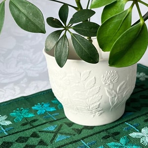 Op de afbeelding: Een witte keramische pot met een reliëfmotief van bloemen, met daarin een groene, bladerige plant met meerdere stengels. De pot staat op een groene placemat met een geometrisch patroon en blauwe borduurwerk.