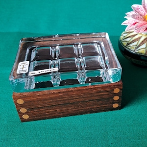 May include: A clear glass ashtray with a wooden base and brass accents. The ashtray has a grid pattern and the text "FULL LEAD CRYSTAL" is printed on the bottom. The ashtray is sitting on a green surface next to a small, white and brown ceramic vase with a pink flower.